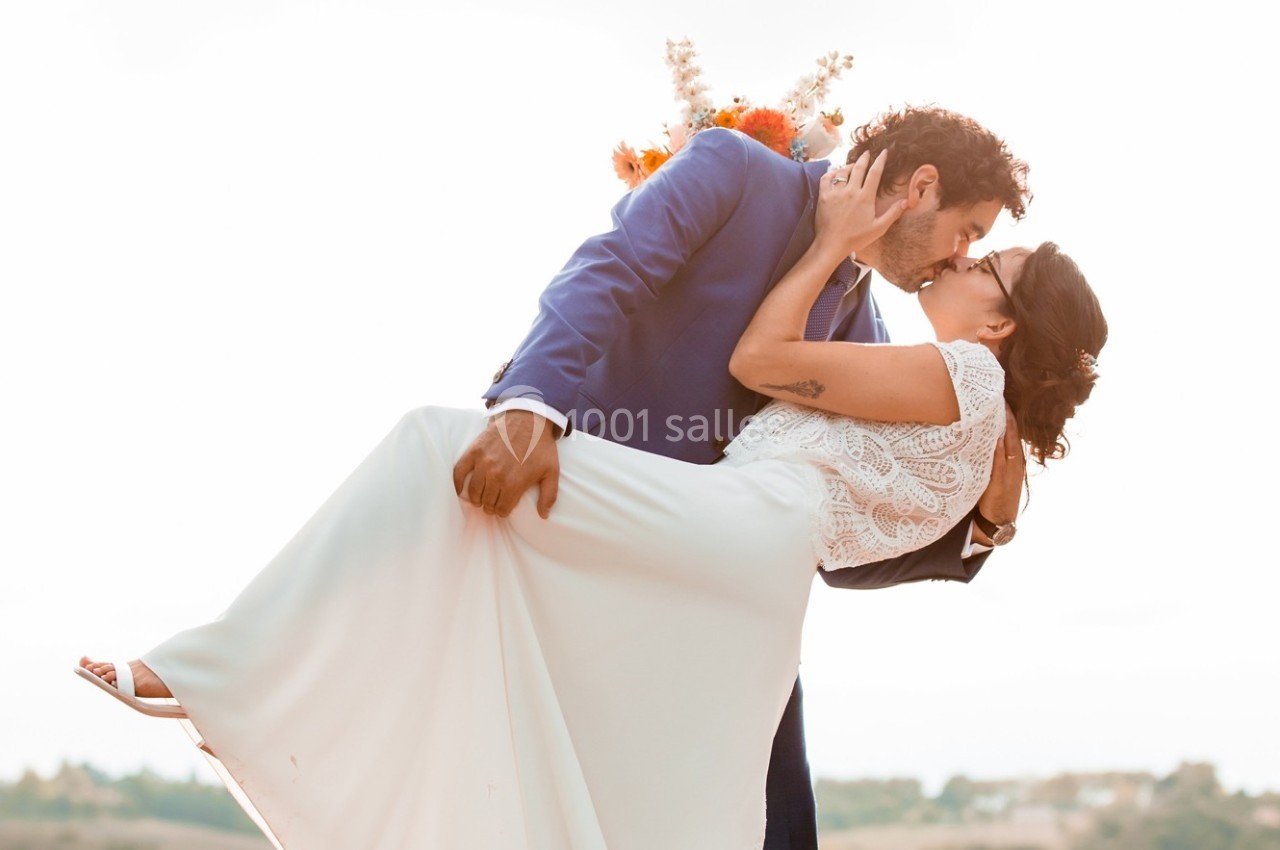 Un couple en tenue de mariage s'embrasse, l'homme portant la femme dans ses bras, avec un bouquet en arrière-plan.