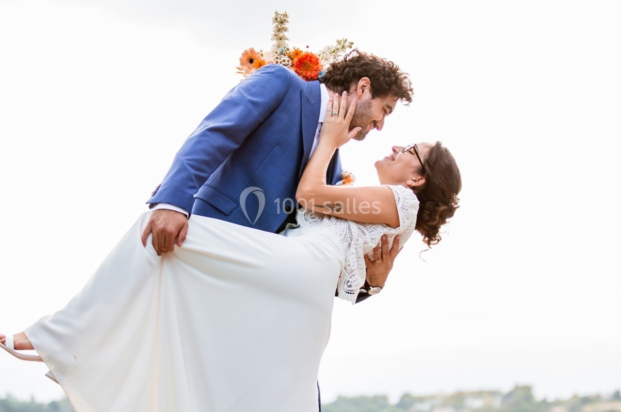 Un homme en costume bleu porte une femme en robe blanche, tous deux souriants dans un cadre extérieur lumineux.