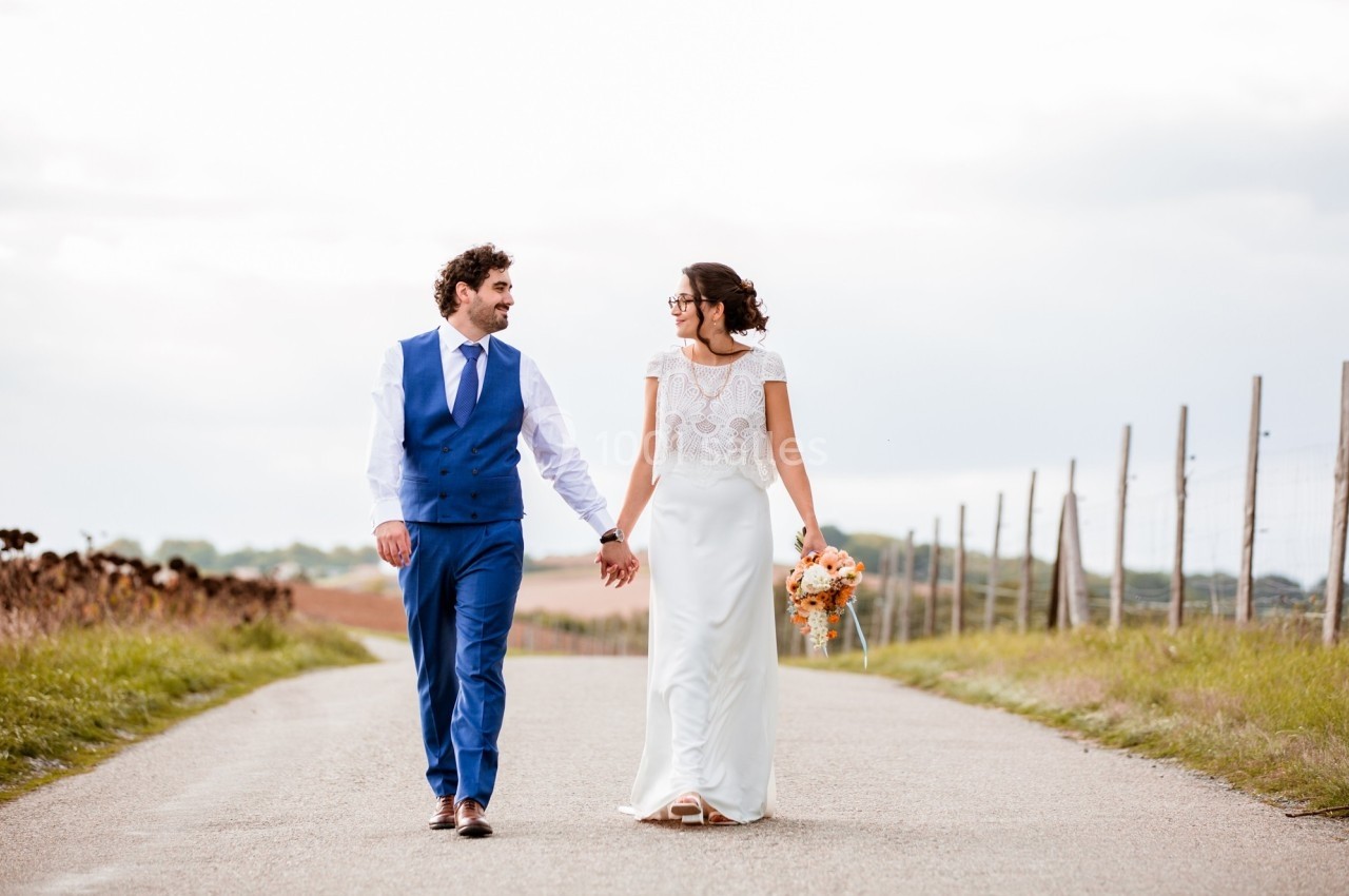 Un couple en tenue de mariage marche main dans la main sur une route de campagne bordée de clôtures.