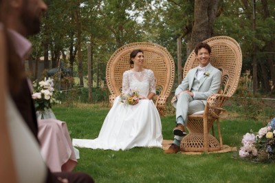 Une mariée en robe blanche tient un bouquet coloré face à un homme en costume clair dans un jardin verdoyant.