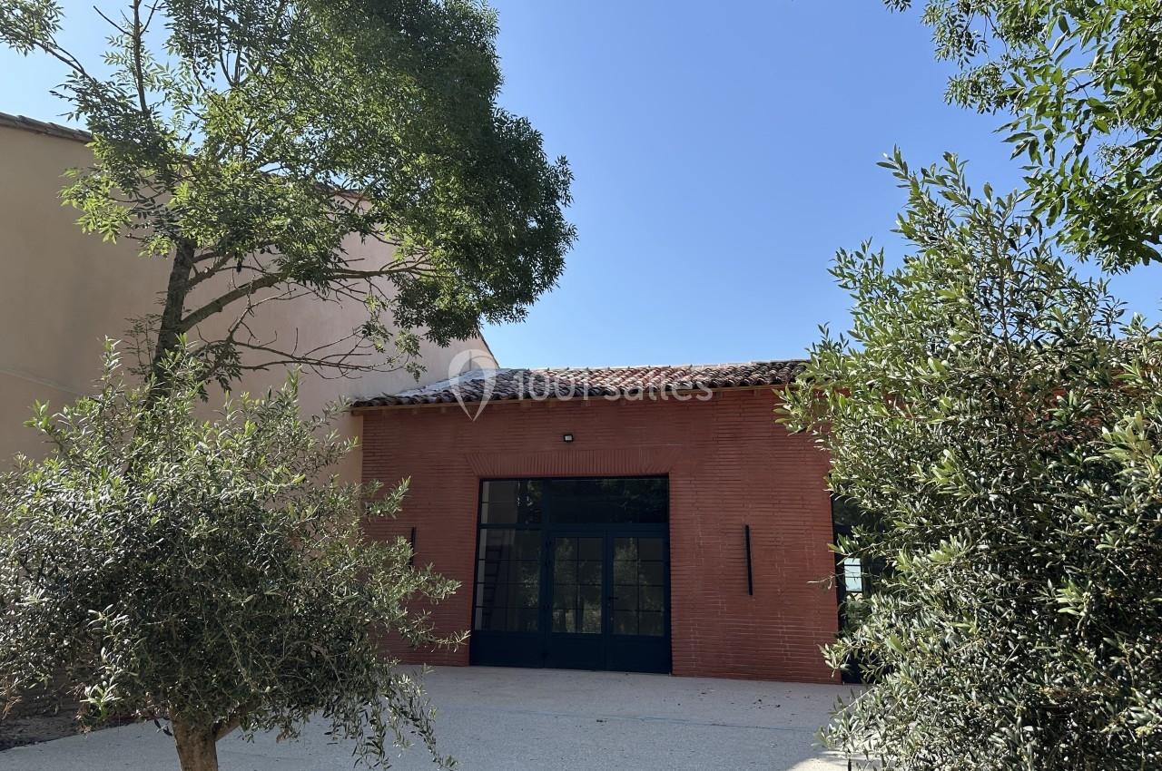 Façade d'un bâtiment en briques rouges avec une grande porte vitrée, entouré d'arbres et d'arbustes sous un ciel bleu.