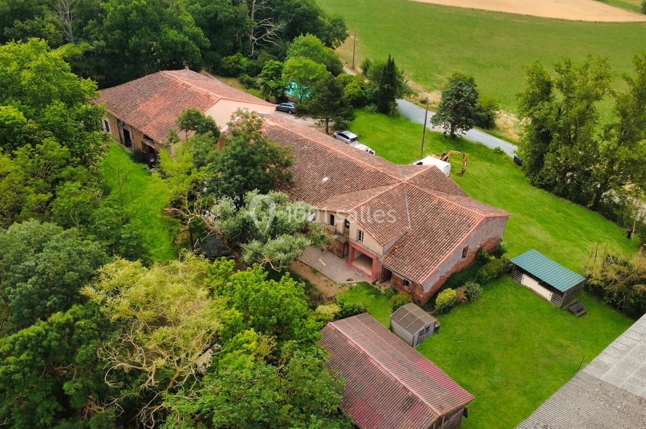 Vue aérienne d'une grande maison en briques avec toit en tuiles, entourée de verdure et de bâtiments annexes.