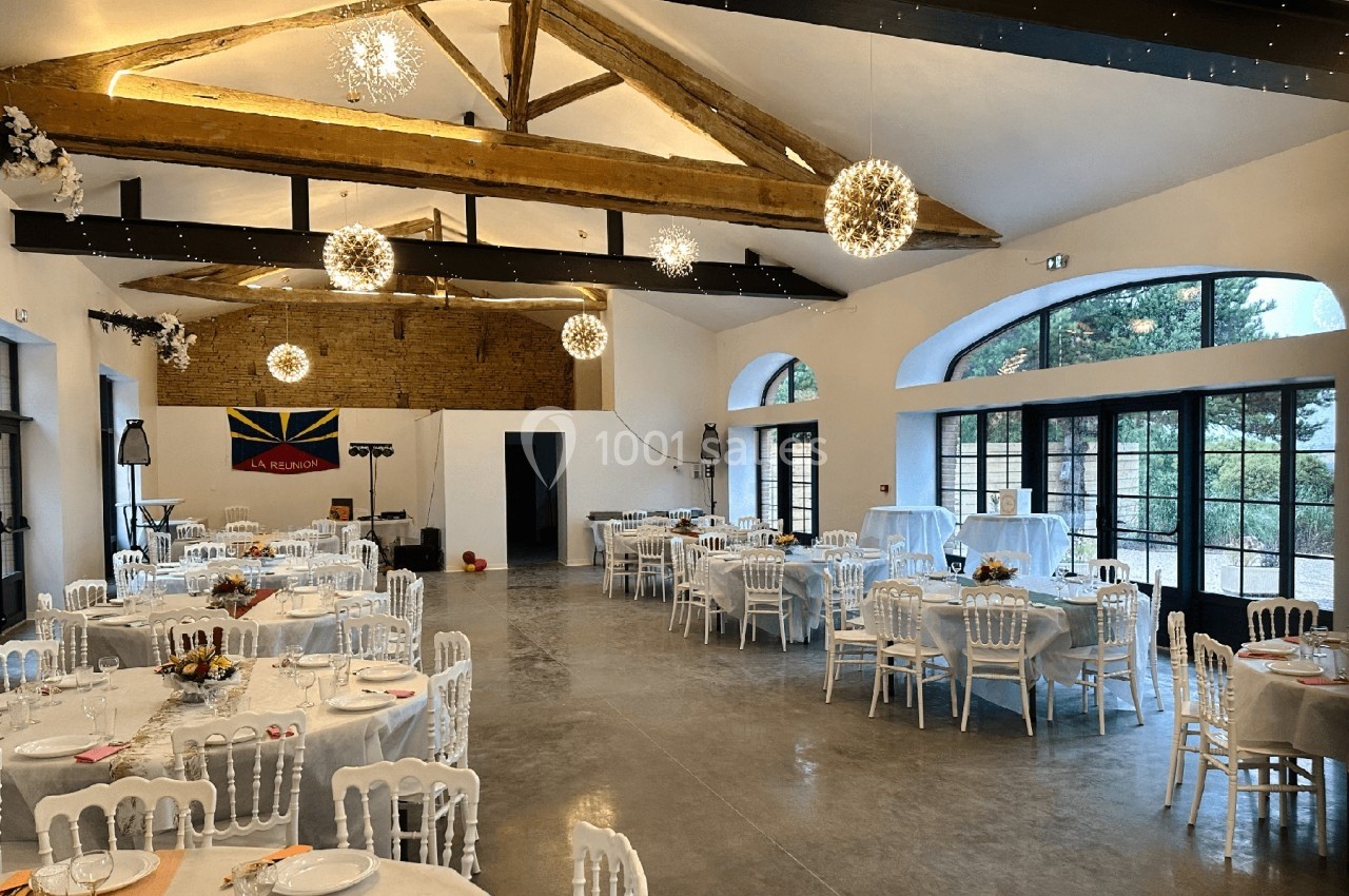 Salle de réception lumineuse avec tables rondes dressées, poutres apparentes et grandes baies vitrées donnant sur l…