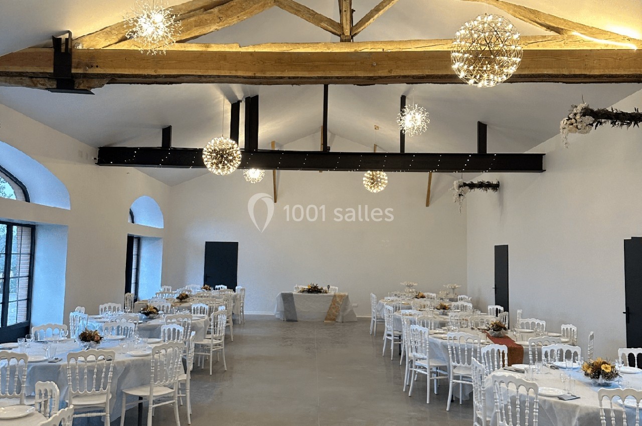 Salle de réception lumineuse avec poutres apparentes, tables rondes dressées et décorations florales.