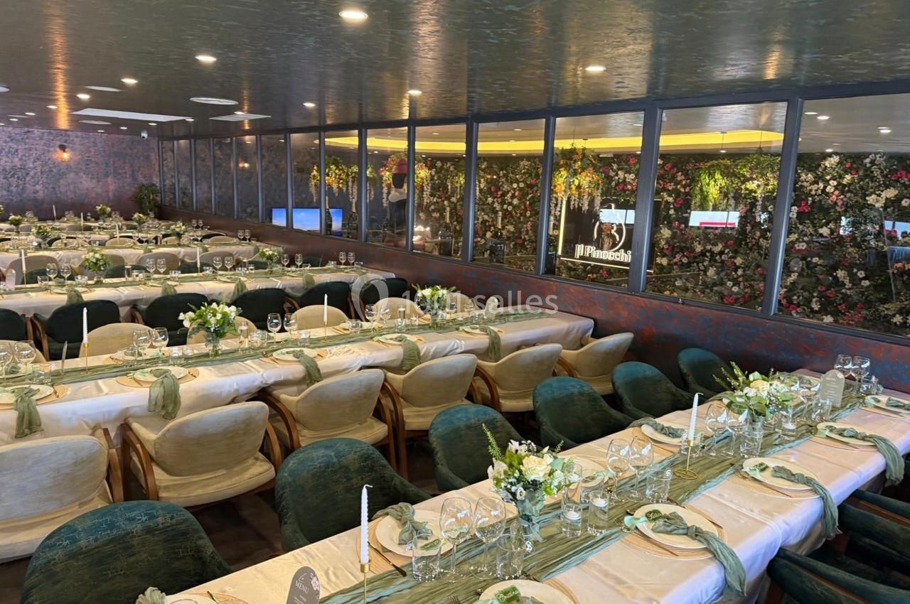 Salle de réception élégamment décorée avec de longues tables dressées, nappes beiges, chaises vertes et arrangements floraux.