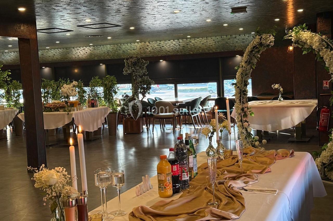 Salle de réception décorée avec des tables rondes, des arches fleuries et une table ornée de bougies et boissons.