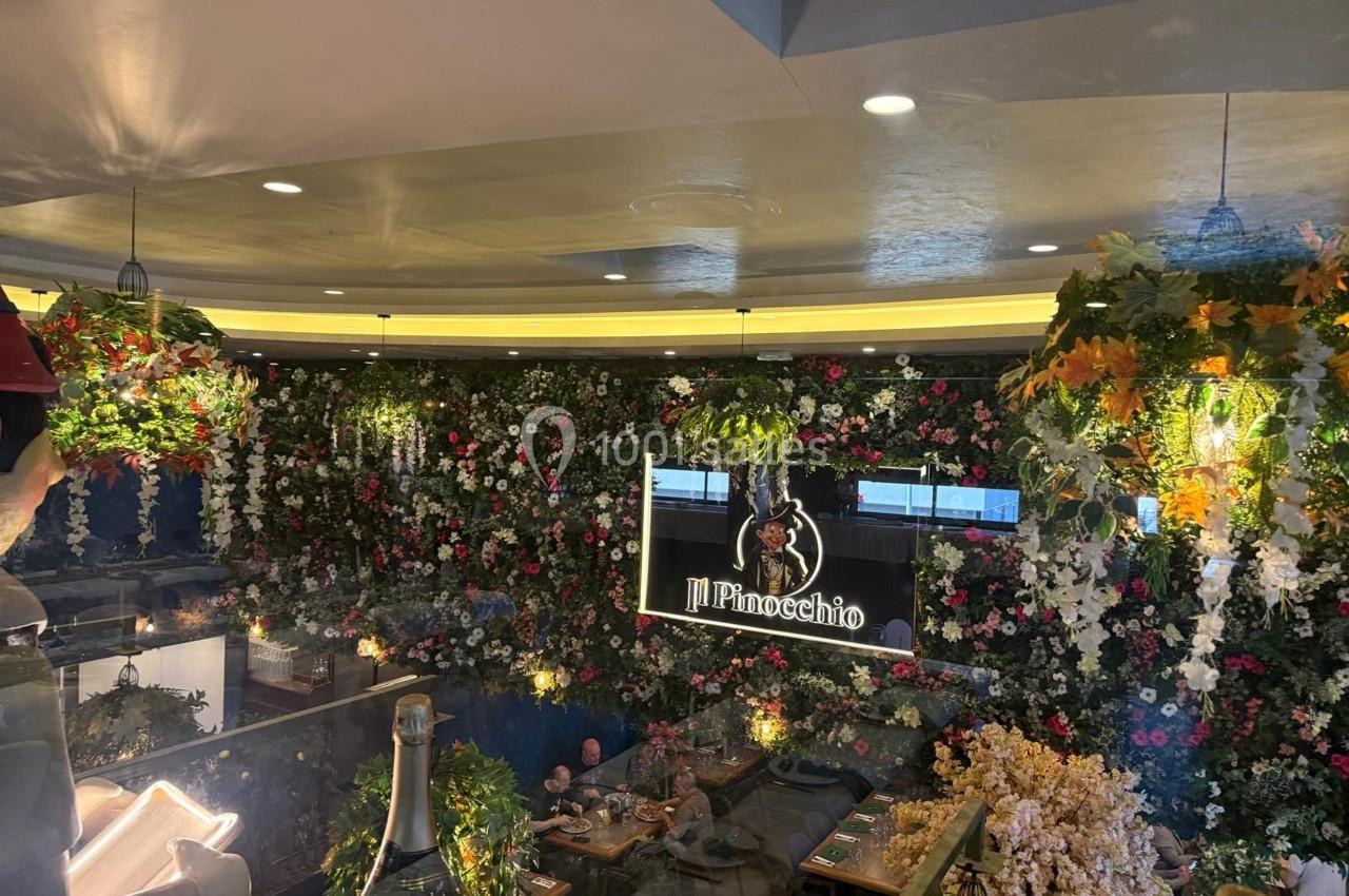 Mur végétal fleuri décorant un restaurant avec des tables dressées et un panneau lumineux ’Il Pinocchio’.