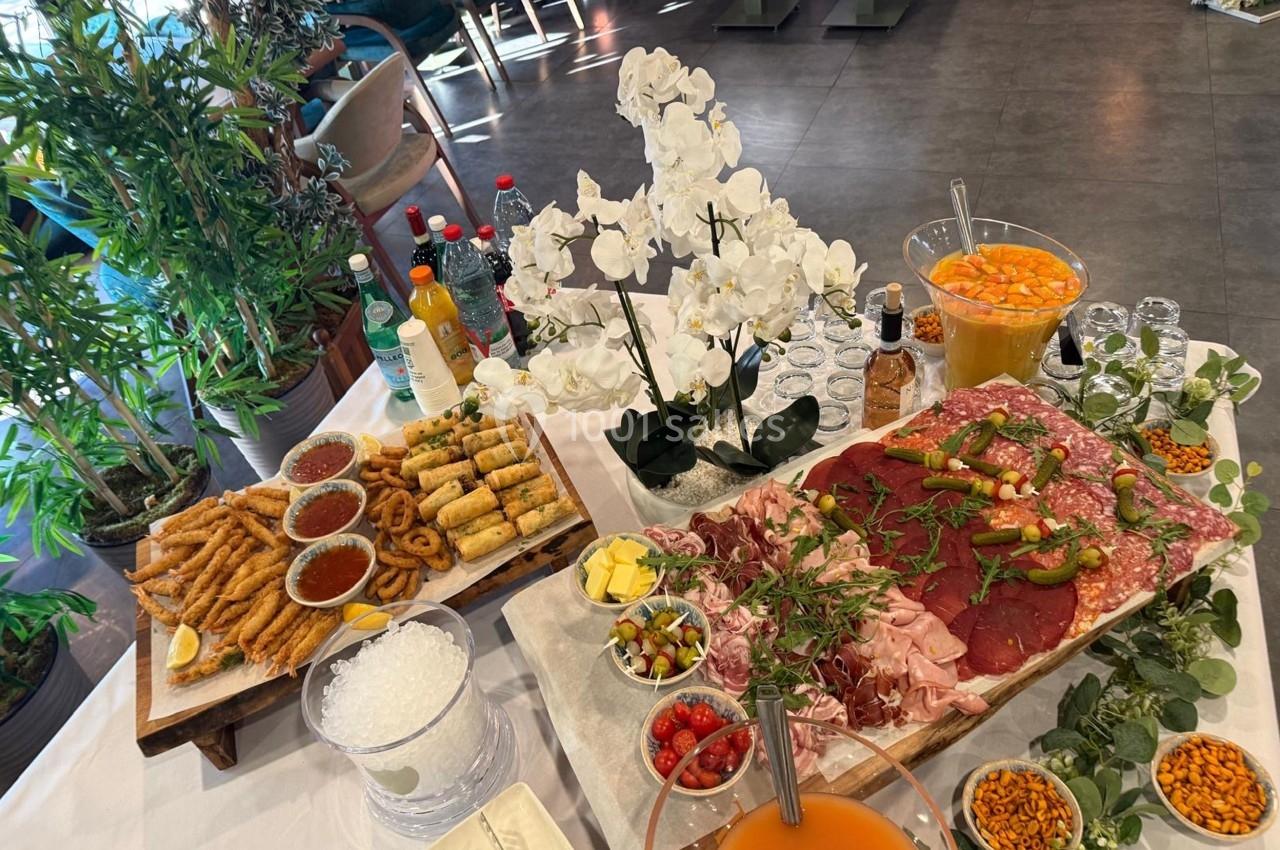 Plateau de charcuterie, beignets, légumes frits et boissons disposés sur une table décorée de fleurs blanches.