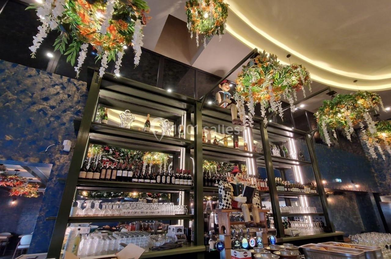 Intérieur d'un bar avec étagères remplies de bouteilles, verres suspendus et décorations florales au plafond.