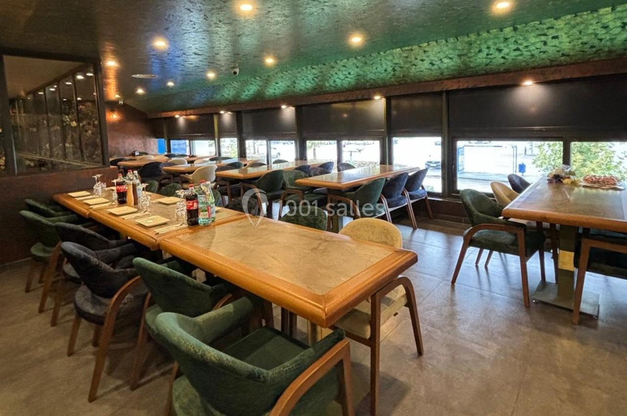 Salle de restaurant avec tables en bois, chaises rembourrées et grandes fenêtres laissant entrer la lumière naturelle.