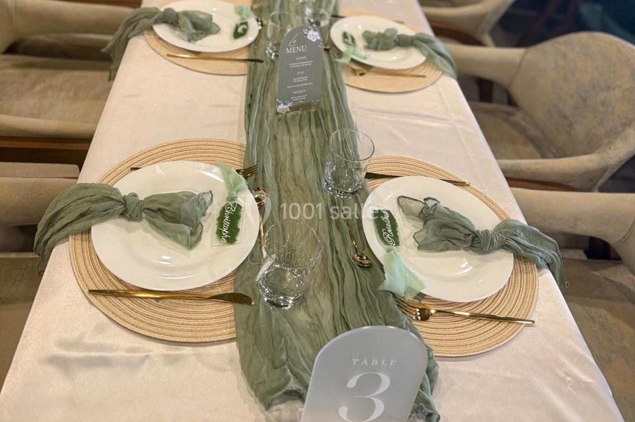 Table dressée avec nappes beige et verte, assiettes blanches, couverts dorés, et décoration élégante en tissu vert.