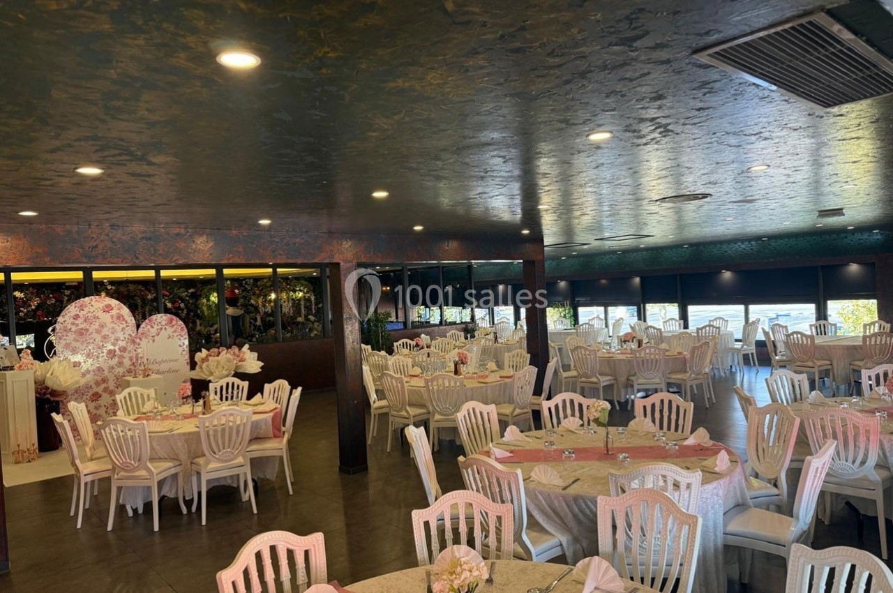 Salle de réception décorée avec des tables rondes dressées, des chaises blanches et un fond floral.