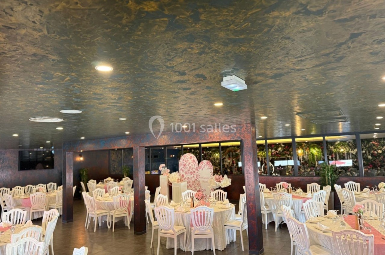 Salle de réception décorée avec des tables rondes dressées, chaises blanches et murs ornés de fleurs.