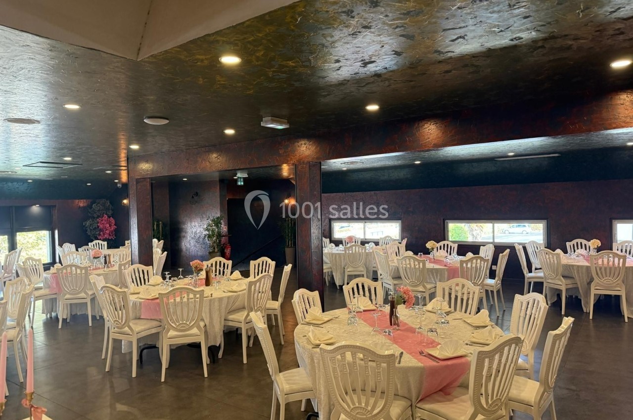 Salle de réception avec des tables rondes dressées, nappes blanches et chaises en plastique blanc, éclairage tamisé.