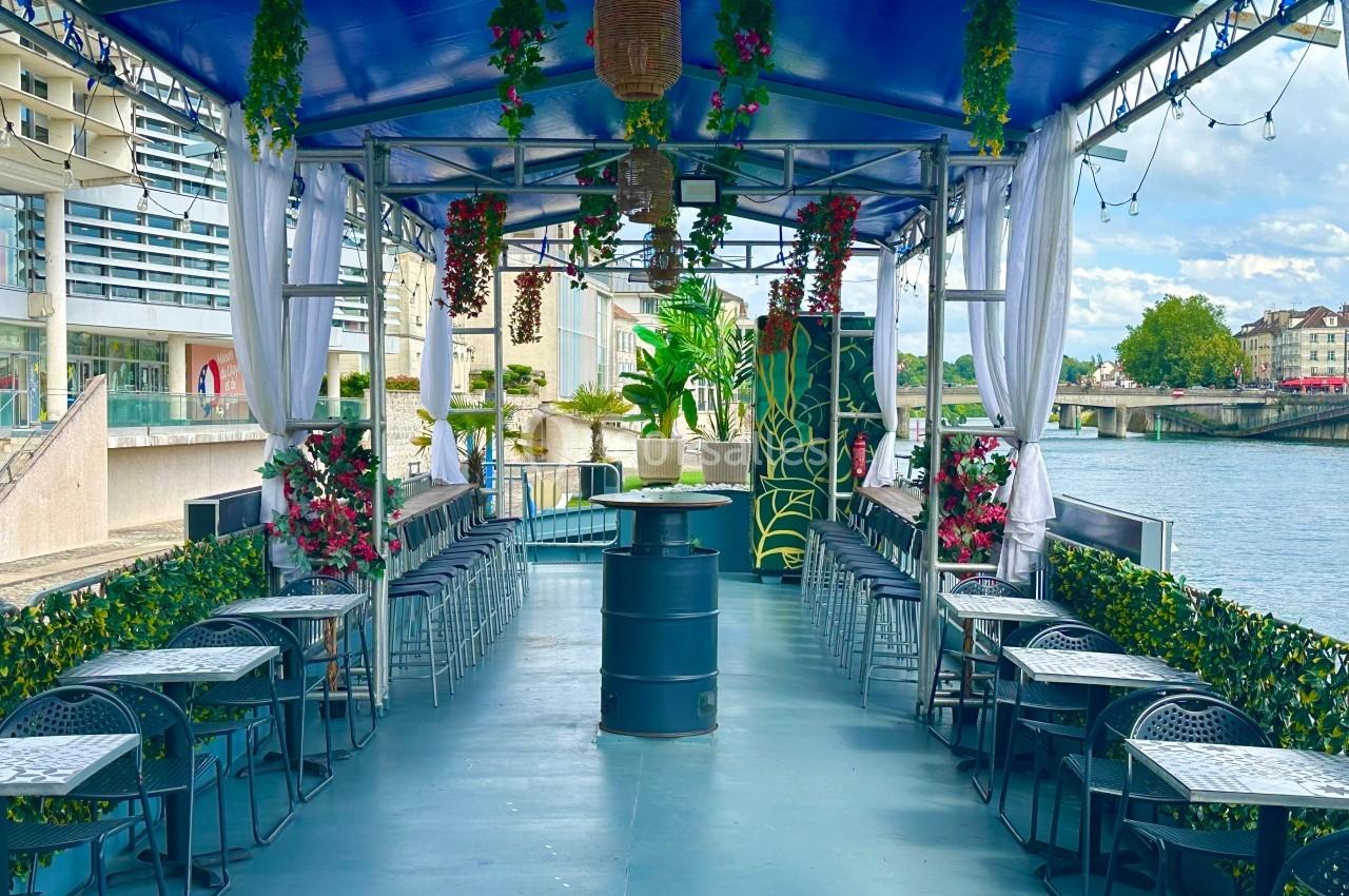 Terrasse couverte sur une péniche, aménagée avec des tables, des chaises et des décorations florales, au bord d'une rivière.