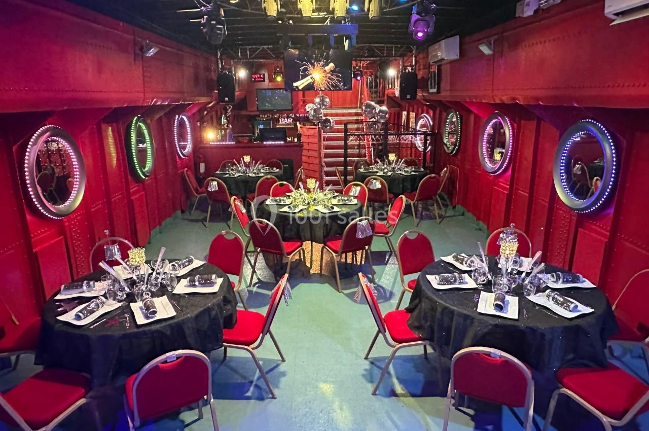 Salle décorée avec des murs rouges, tables rondes dressées pour un repas, éclairage tamisé et ambiance festive.