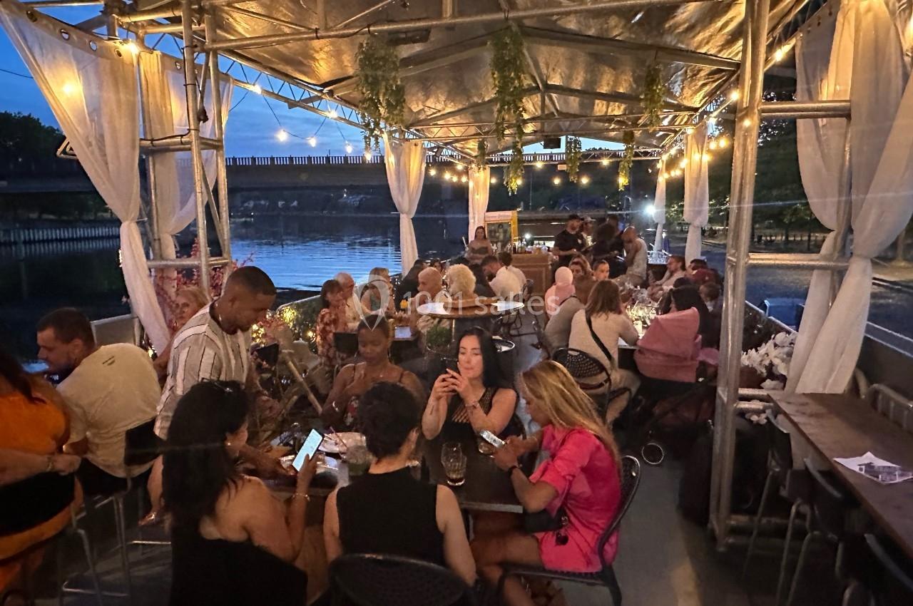 Des personnes dînent et discutent sur une terrasse éclairée en soirée, située au bord de l'eau.