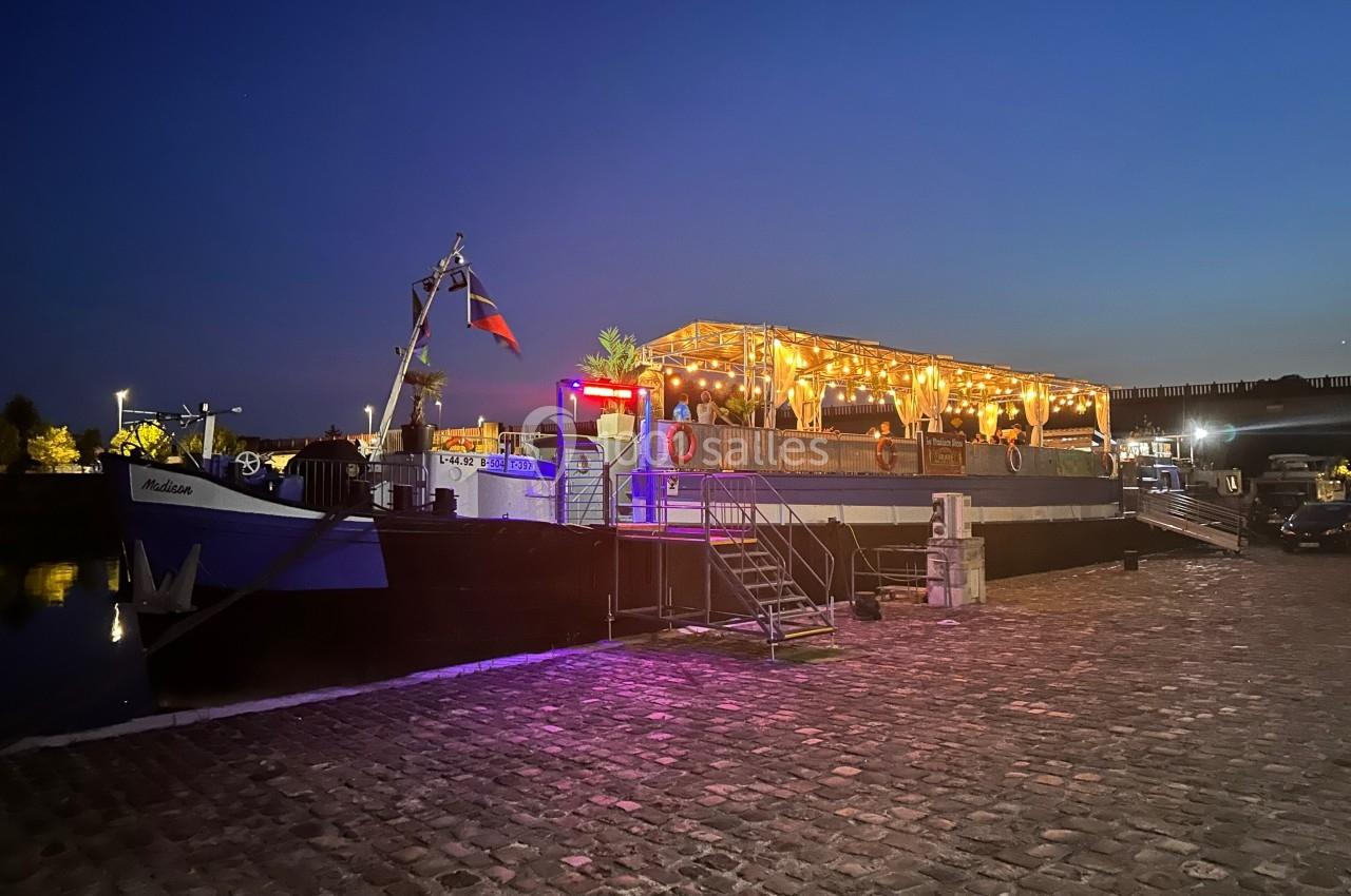 Bateau illuminé amarré le long d'un quai pavé au crépuscule, avec des lumières chaleureuses et des décorations.