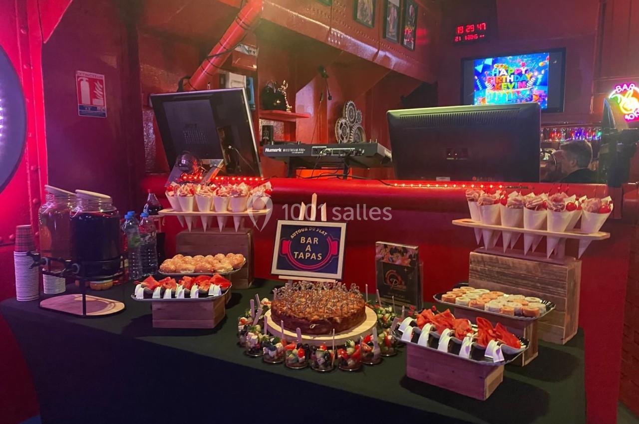 Table garnie de tapas variés, desserts et boissons, dans un bar à l'ambiance lumineuse et festive.