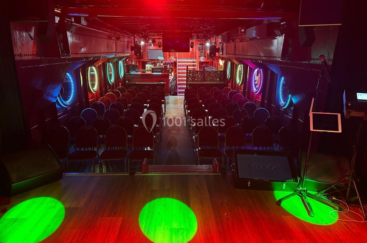 Salle de spectacle avec rangées de sièges, éclairage coloré et scène en bois au premier plan.