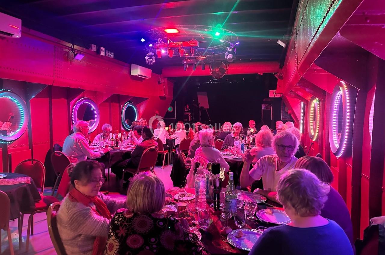 Des personnes dînent dans une salle éclairée par des lumières colorées, avec des hublots lumineux sur les murs rouges.