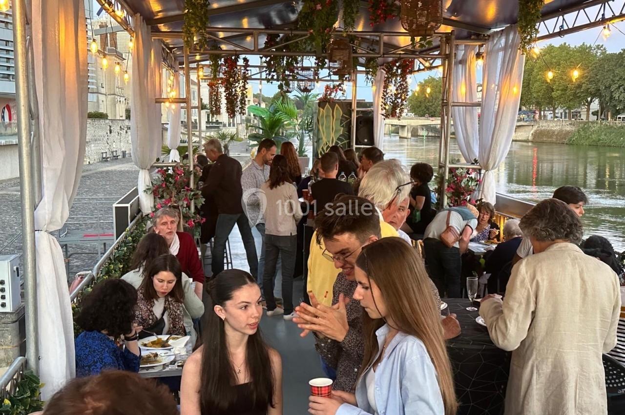 Personnes discutant et partageant un moment convivial sur une péniche décorée, amarrée au bord d'une rivière.