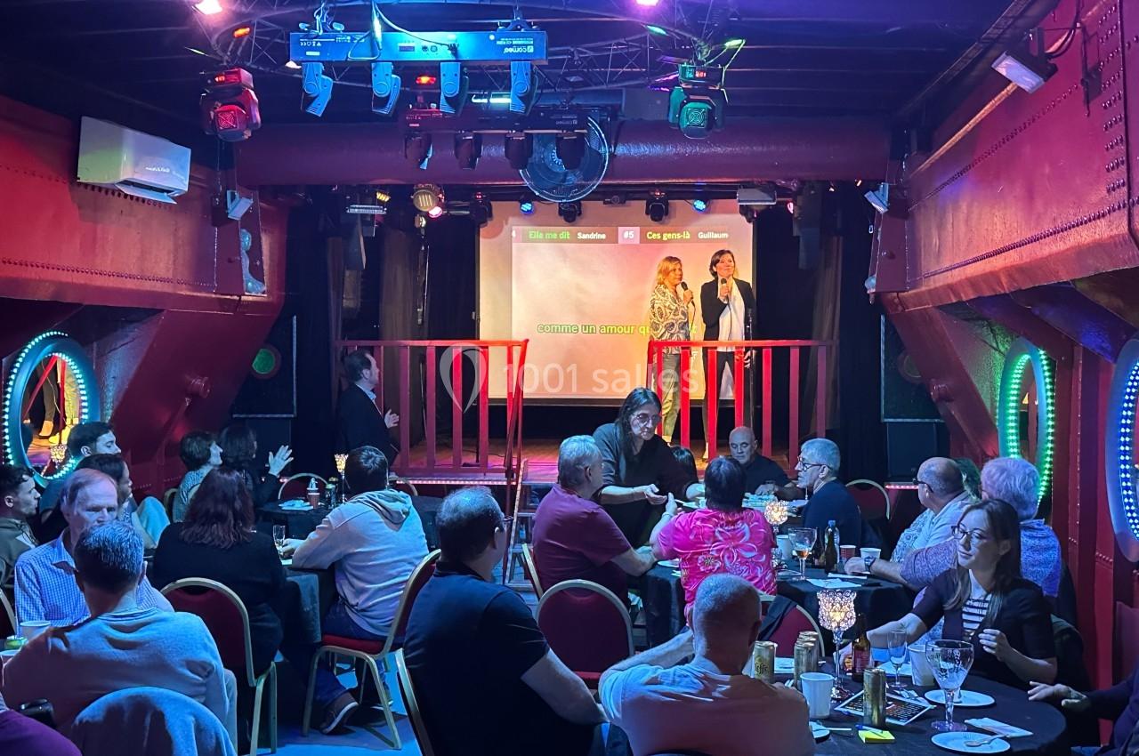Personnes assises à des tables dans une salle, regardant une scène éclairée où deux individus parlent devant un écran.