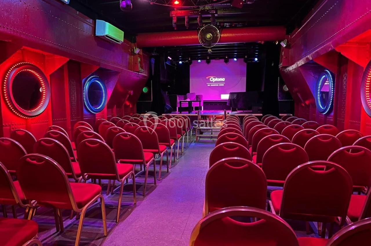 Salle de spectacle avec des rangées de chaises rouges, éclairage coloré et écran de projection en fond.