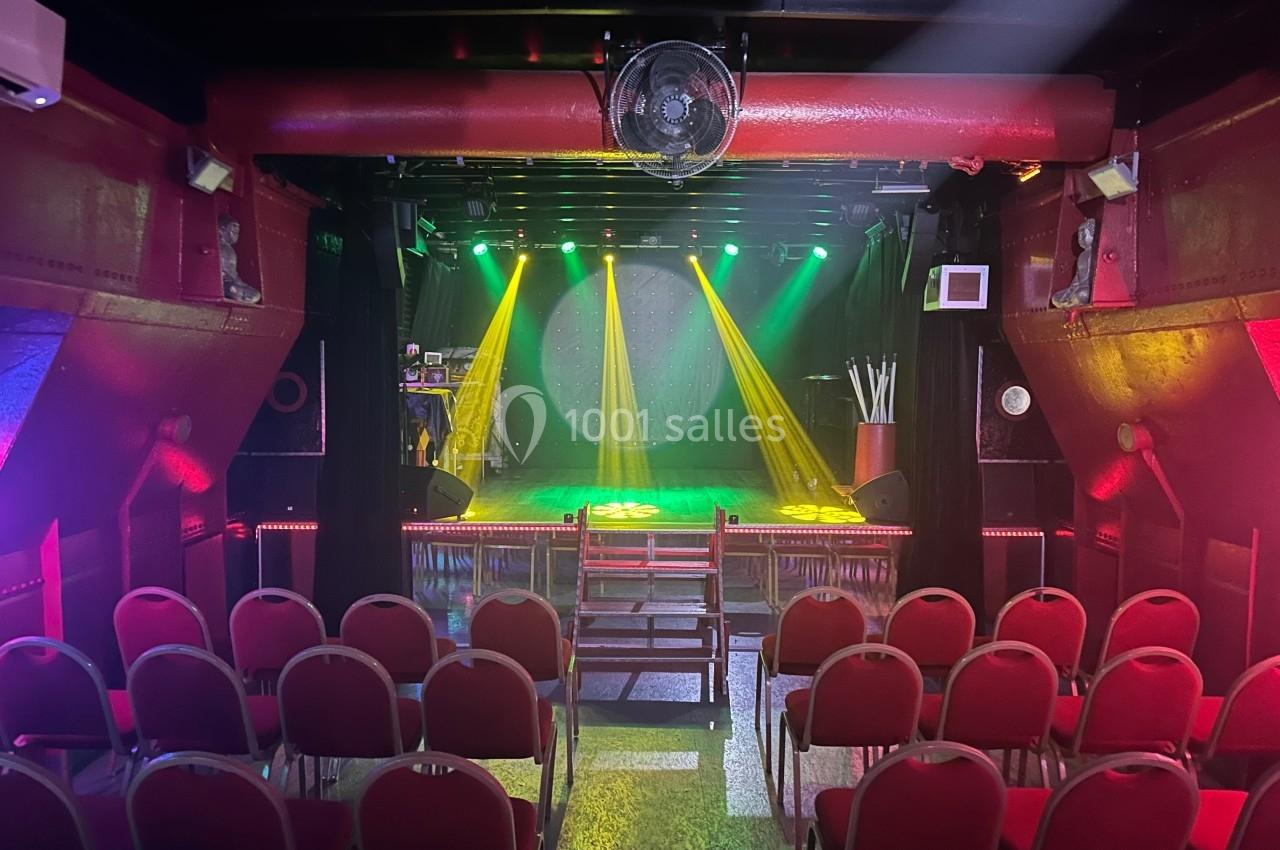 Salle de spectacle avec sièges rouges, scène éclairée par des projecteurs colorés et ambiance tamisée.