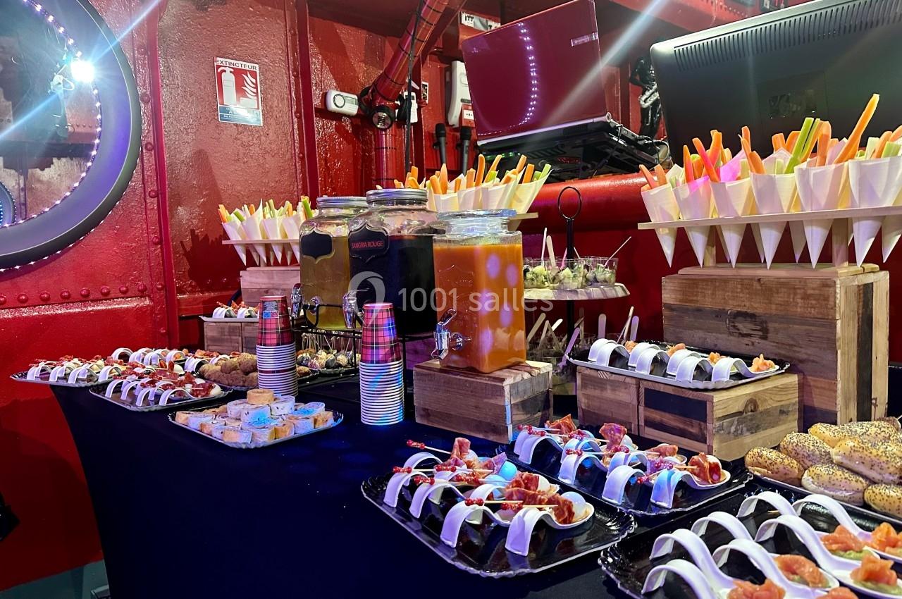 Buffet coloré avec verrines, amuse-bouches, jus et snacks disposés sur une table dans un décor rouge industriel.