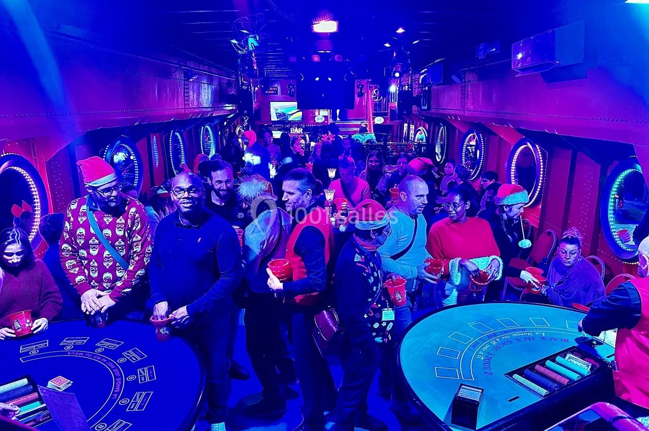 Salle animée avec des tables de jeu, des personnes en tenue festive et une ambiance lumineuse bleutée.
