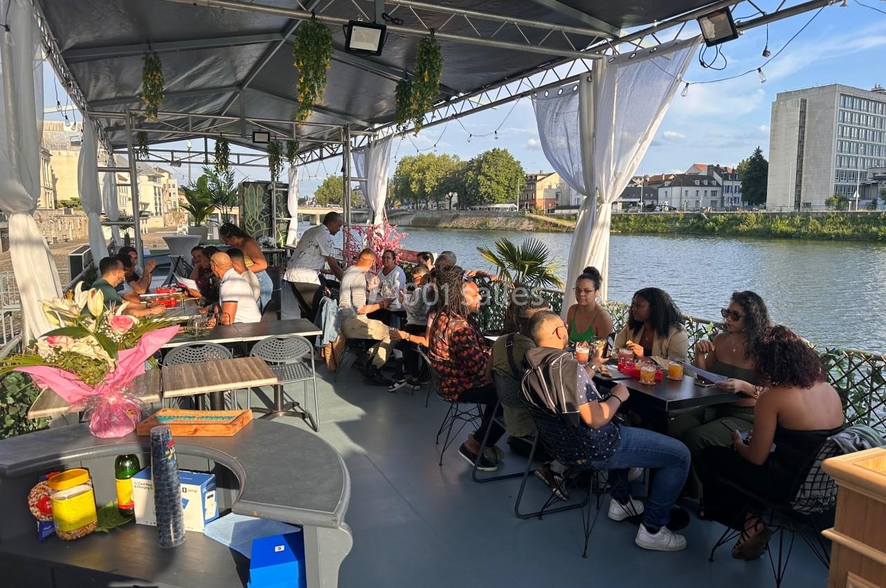 Des personnes dînent et discutent sur une terrasse couverte au bord d'une rivière, par une journée ensoleillée.