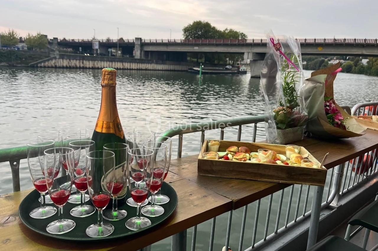 Bouteille de champagne, coupes et amuse-bouches disposés sur une table en bois, avec vue sur une rivière et un pont.