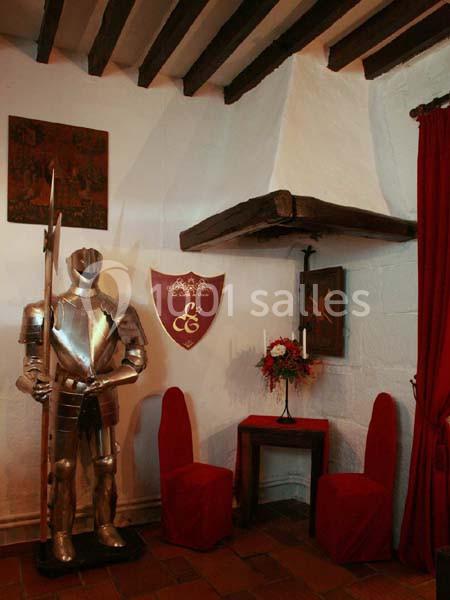 Armure médiévale exposée dans une pièce décorée avec des chaises rouges, un blason et des poutres en bois.