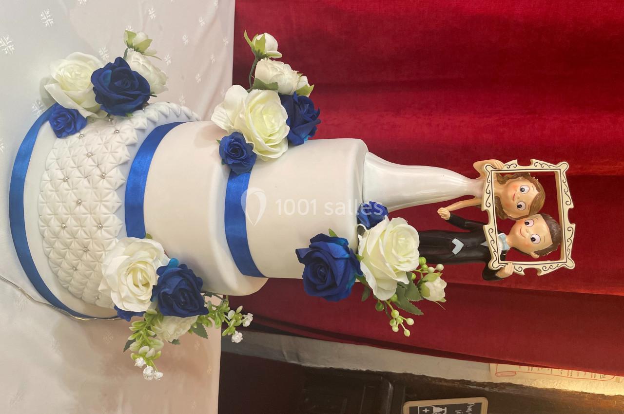 Gâteau de mariage blanc décoré de roses bleues et blanches, avec des figurines humoristiques en haut.