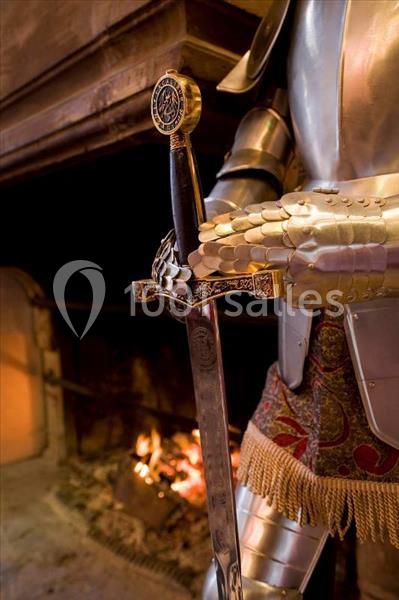 Armure médiévale tenant une épée décorée, devant une cheminée allumée dans un intérieur chaleureux.