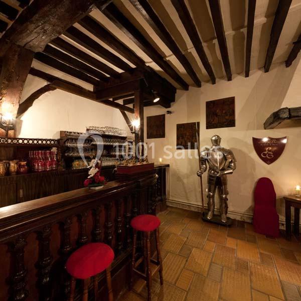 Salle rustique avec poutres apparentes, bar en bois, tabourets rouges, armure médiévale et bougies décoratives.