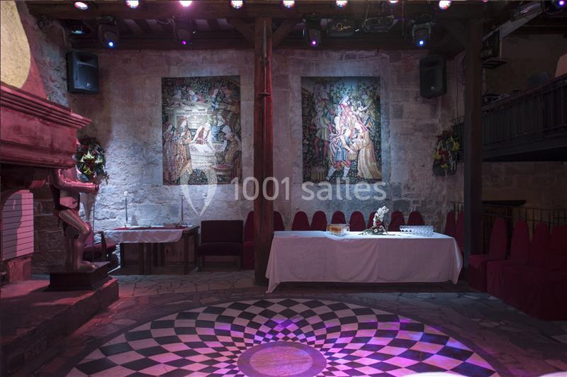 Salle médiévale avec tapisseries murales, cheminée en pierre, table dressée et sol à motif géométrique éclairé.