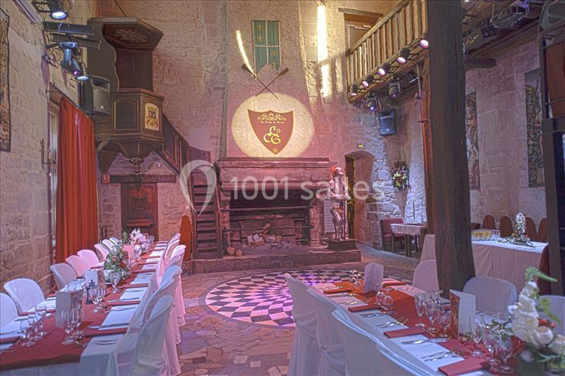 Salle médiévale avec cheminée en pierre, tables dressées pour un repas, et décorations historiques sur les murs.