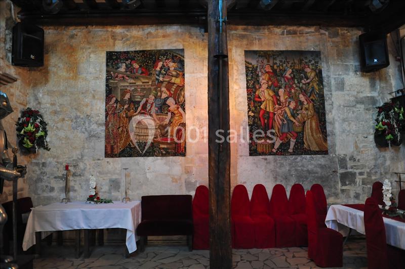 Deux tapisseries médiévales décorent un mur en pierre dans une salle avec des chaises rouges et des tables dressées.