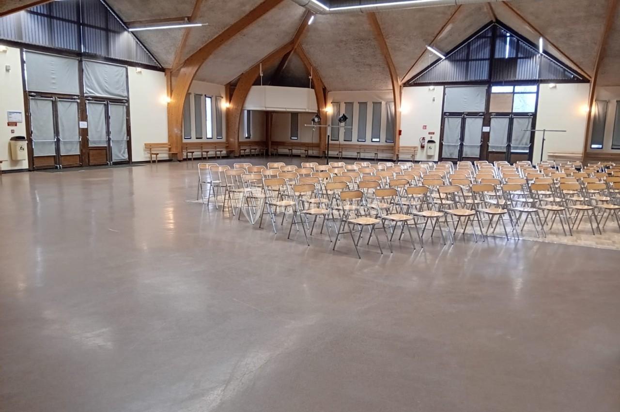 Salle spacieuse avec des chaises alignées, plafond en bois et grandes fenêtres laissant entrer la lumière naturelle.