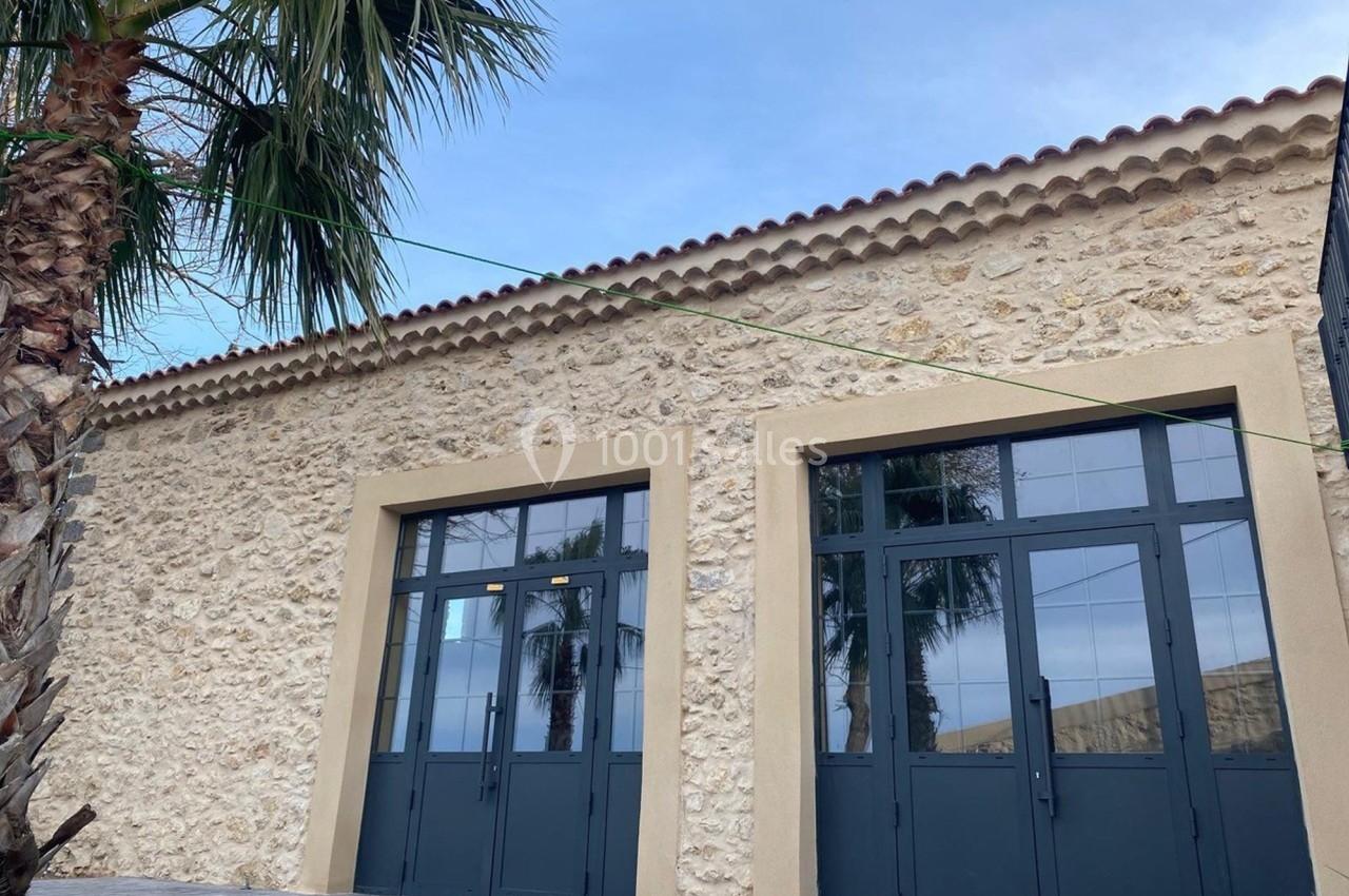 Façade en pierre avec deux grandes portes vitrées noires, entourée de palmiers sous un ciel légèrement nuageux.