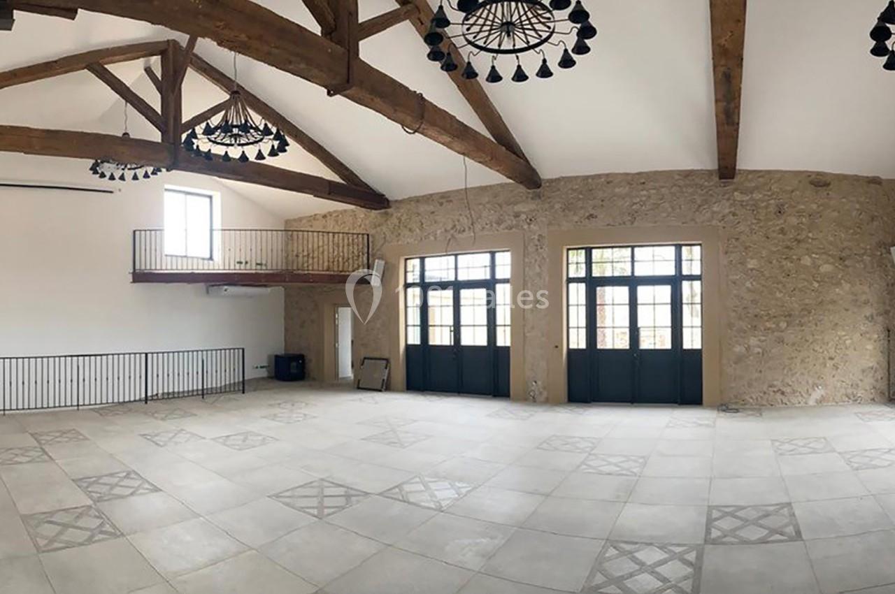 Salle spacieuse avec poutres en bois apparentes, murs en pierre, grandes fenêtres et mezzanine métallique.