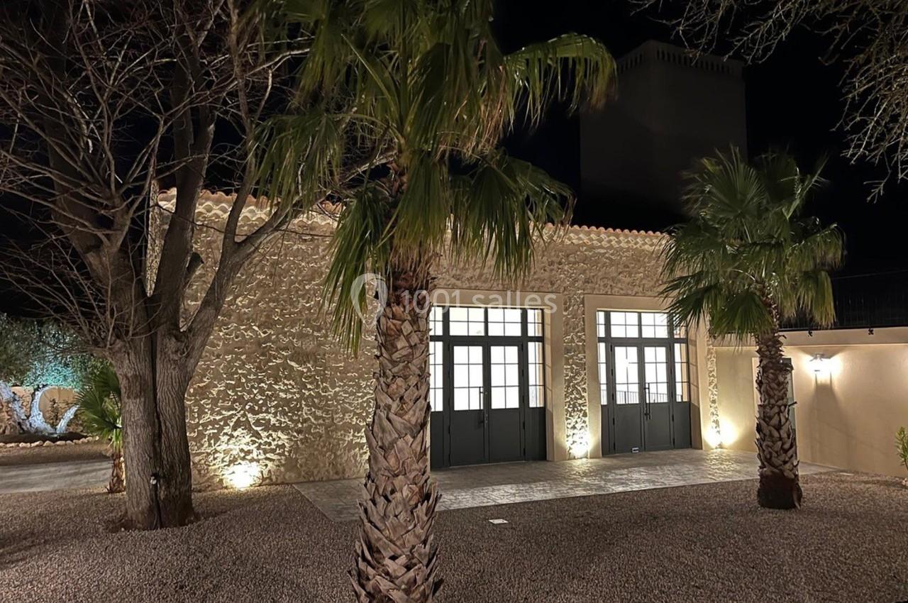 Façade d'un bâtiment en pierre éclairé de nuit, entouré de palmiers et d'arbres sur un sol gravillonné.