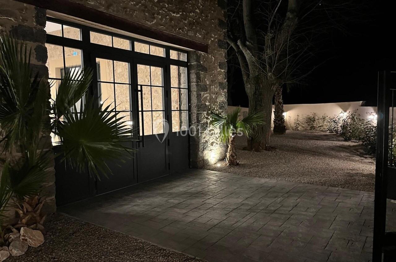 Entrée vitrée d'un bâtiment en pierre, éclairée la nuit, avec des arbres et des plantes dans une cour pavée.