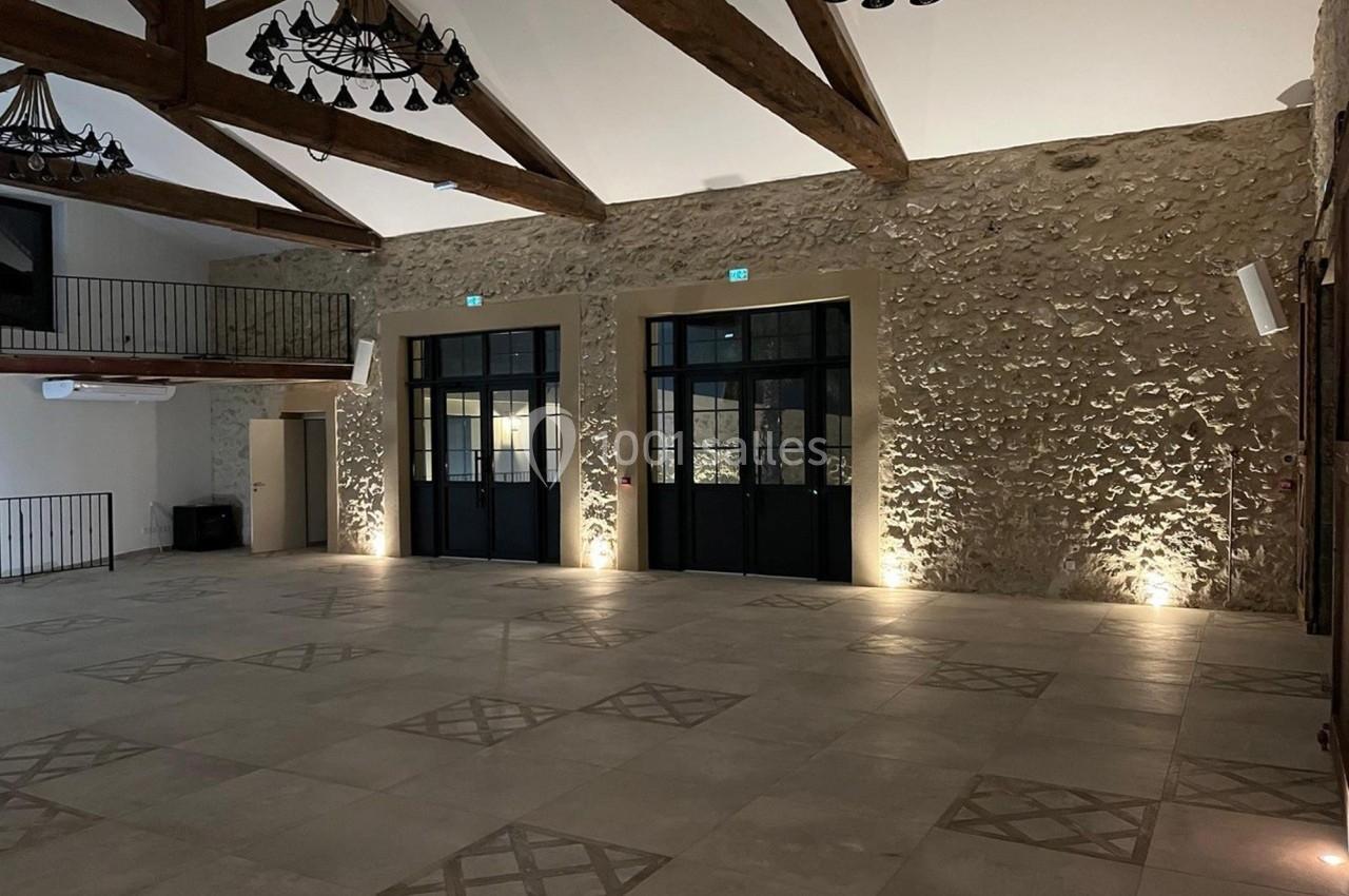 Grande salle avec murs en pierre, poutres apparentes, lustres suspendus et sol en carreaux décoratifs.