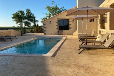 Terrasse en pierre avec piscine, chaises longues, parasols et bâtiments en pierre sous un ciel dégagé.