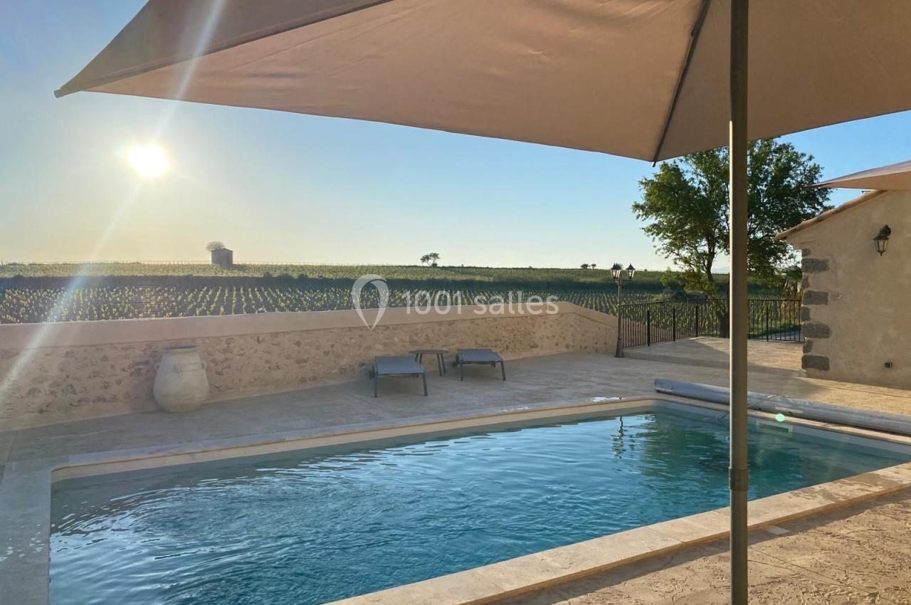 Piscine extérieure entourée de pierres, avec vue sur un champ ensoleillé et un parasol au premier plan.