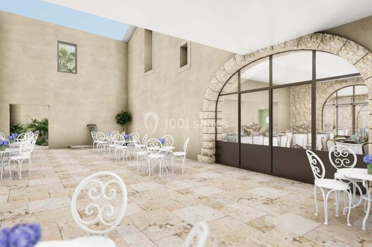 Terrasse en pierre avec tables et chaises blanches, décorée de fleurs violettes, entourée de murs beige et grandes baies…