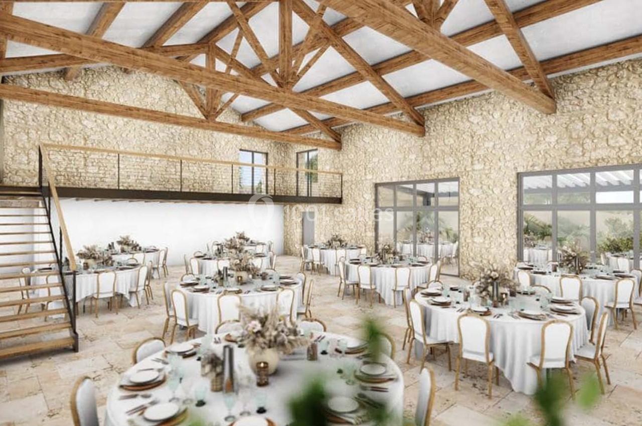 Salle de réception lumineuse avec murs en pierre, poutres apparentes, tables rondes dressées et mezzanine en bois.